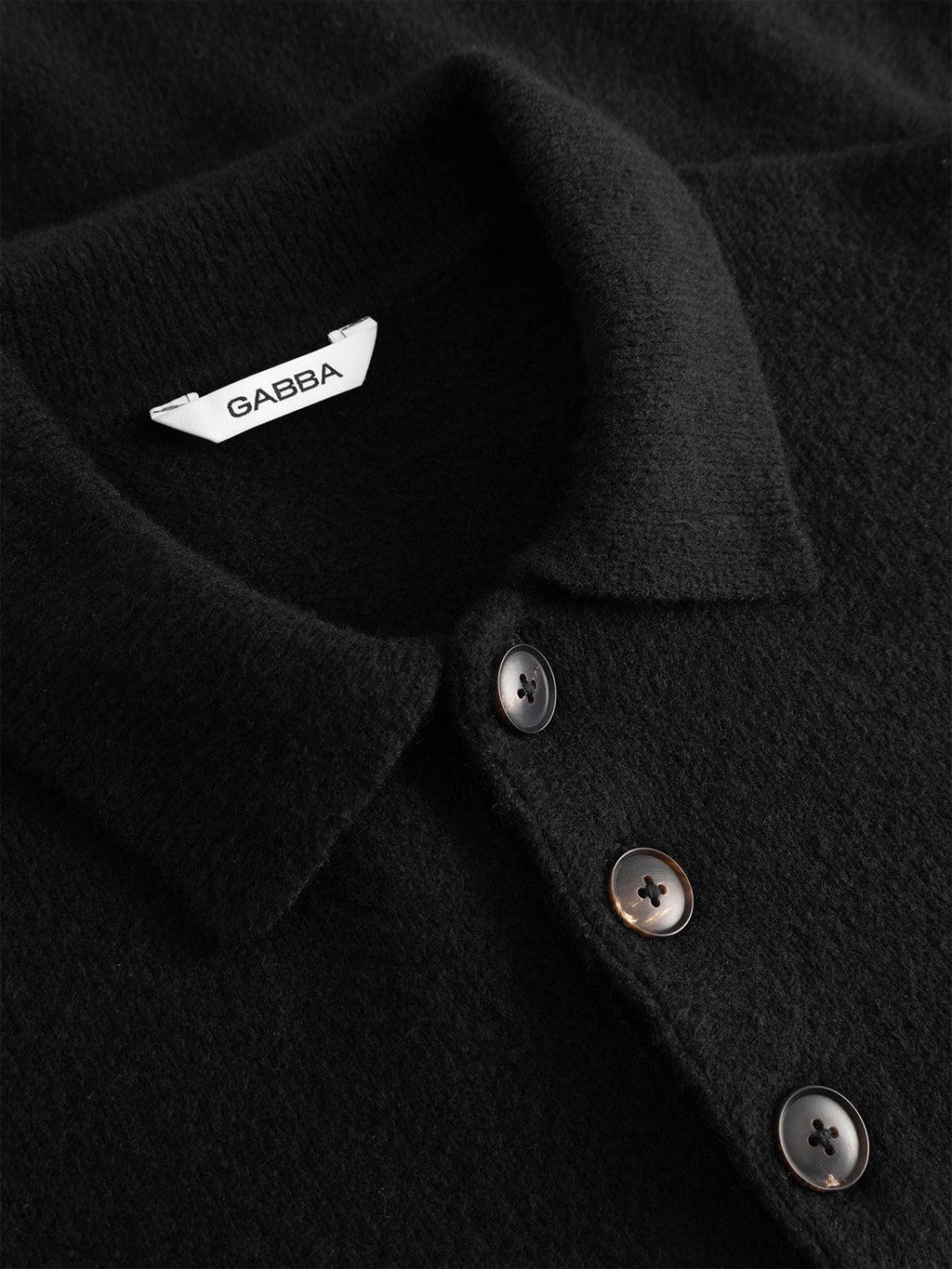 Close-up of the GABpietro Polo Argyle LS Knit in black by Gabba—a mid-weight cotton blend polo shirt featuring a collar, three dark buttons, and a white Gabba-labeled clothing tag.
