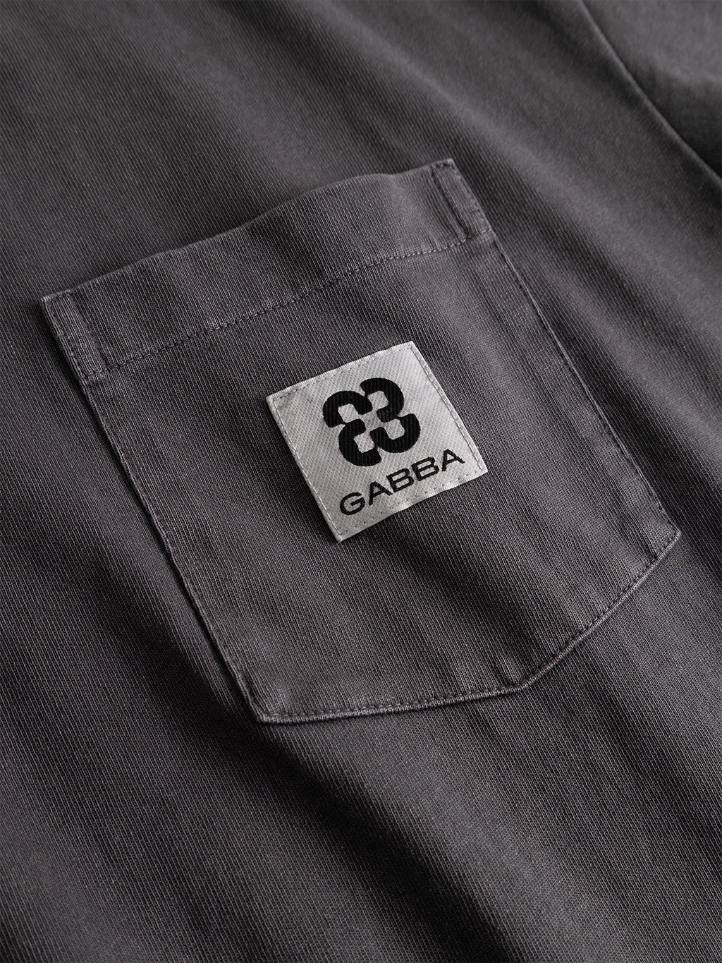 A close-up of the Gabba GABnigel Wix Pkt Boxy Hour SS T-Shirt in dark grey, made from heavyweight cotton with a boxy fit, featuring a chest pocket adorned with a white rectangular patch above the opening that displays a black abstract logo and the bold word “GABBA.”.