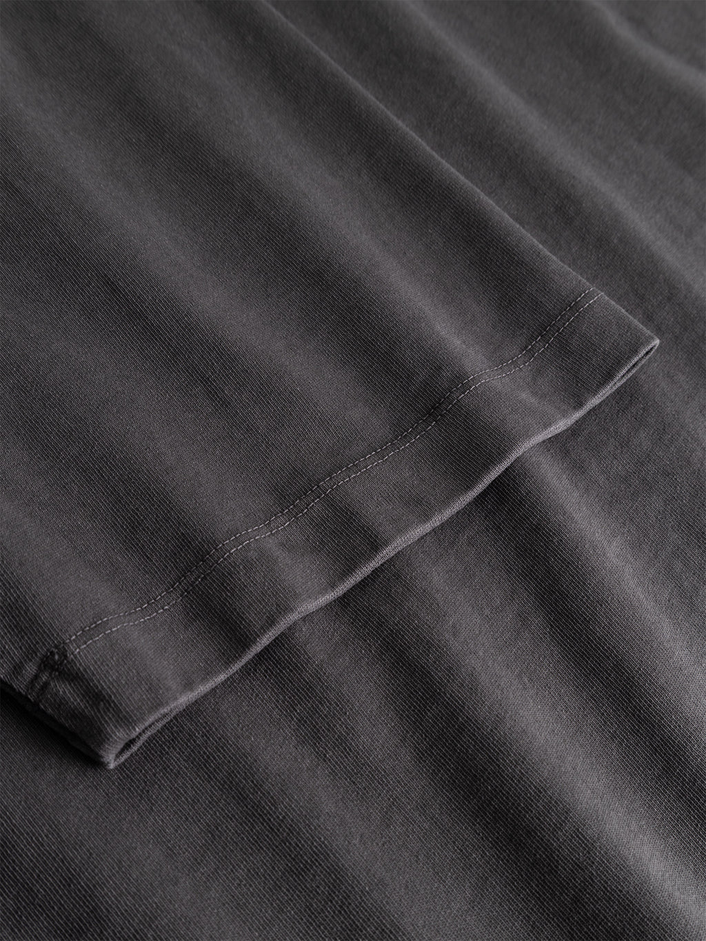 Close-up view of the hem and stitching on the sleeve of the Gabba GABnigel Wix Pkt Boxy Hour SS T-Shirt in dark grey, made from heavyweight cotton and laid flat on a matching surface.