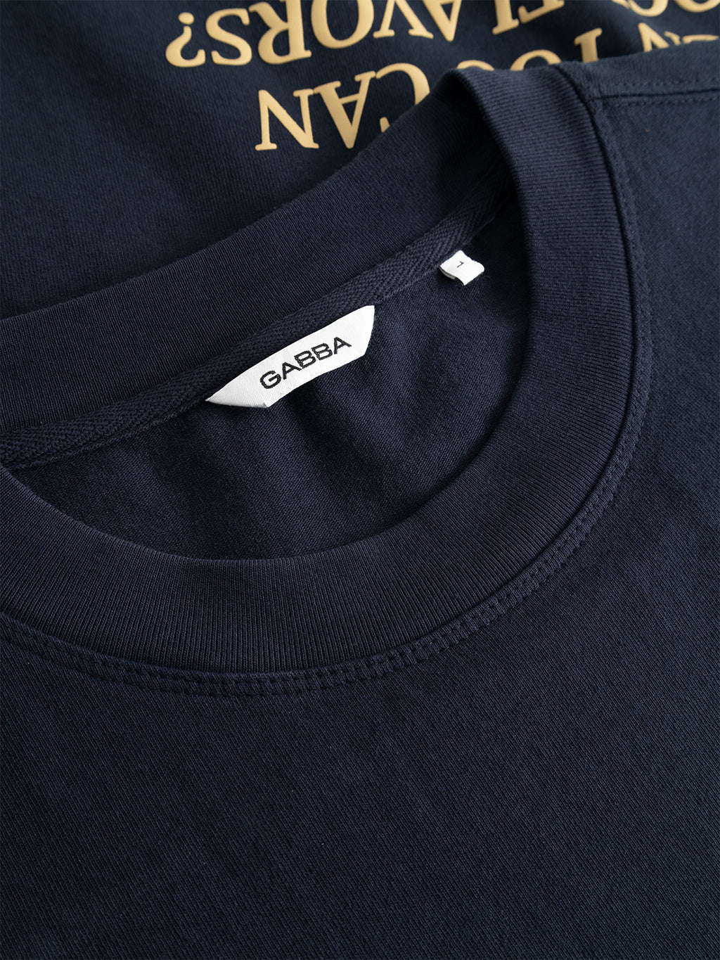 Close-up of the collar of the Gabba GABlegin Wix Boxy Hour Lobby2 SS T-shirt in Night Sky, featuring a Gabba label and a size small tag on textured cotton jersey. Another dark shirt with gold text appears partially underneath.