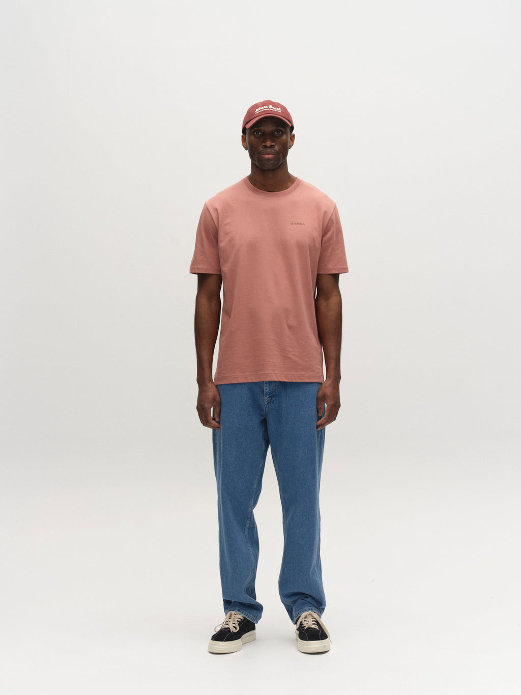 A man faces forward against a plain white background, wearing the Gabba GABdune Logo GOTS SS T-Shirt in Wood Pink, blue jeans, black sneakers with white soles, and a matching pink baseball cap.