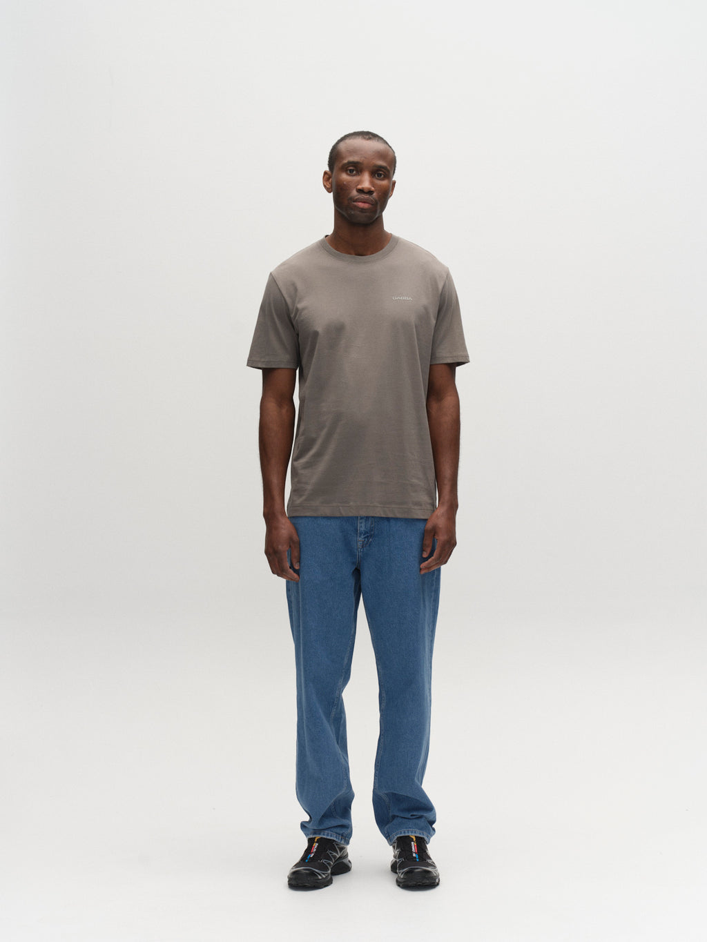 A man stands facing forward against a white background, wearing the Gabba GABdune Logo GOTS SS T-Shirt in Castor Gray, blue jeans, and black shoes. His arms are relaxed by his sides and he has a neutral expression.