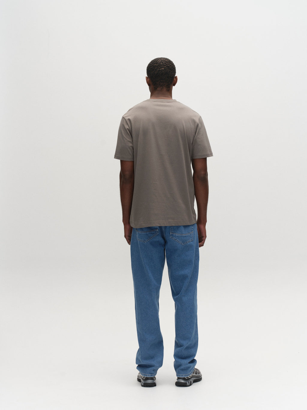 A person stands facing away from the camera against a plain white background, wearing the Gabba GABdune Logo GOTS SS T-Shirt in Castor Gray, blue jeans, and black sneakers.