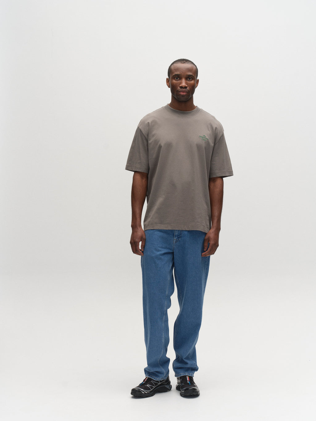 A man stands against a white background, wearing the Gabba GABnigel Boxy NBH SS T-shirt GOTS in Castor Gray, paired with blue jeans and black sneakers. He faces forward with a neutral expression, arms relaxed at his sides.