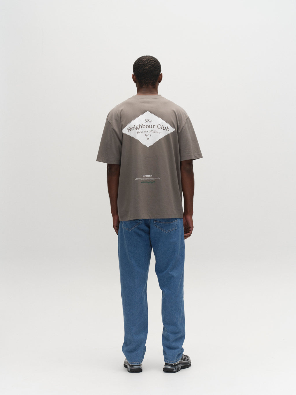 A person stands facing away, wearing the Gabba GABnigel Boxy NBH SS T-shirt GOTS in Castor Gray with “The Neighbour Club” print on the back, blue jeans, and sneakers, against a plain white background.