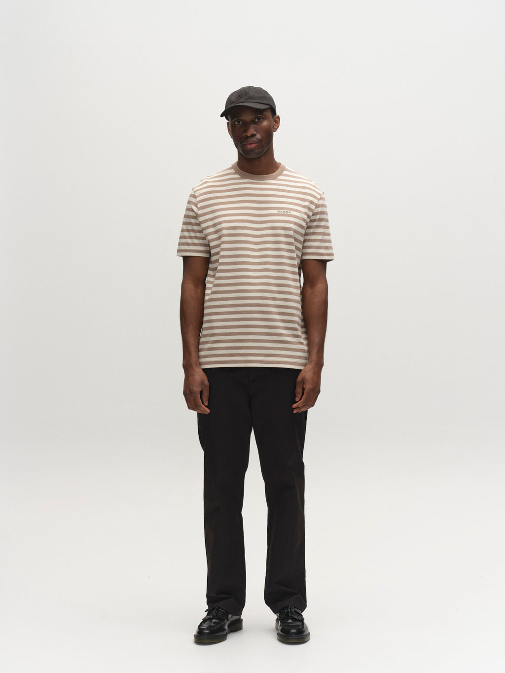 A man stands against a white background, wearing a black cap, black pants, black shoes, and the Gabba GABdune Logo Stripes SS GOTS - Fungi beige and white striped organic cotton t-shirt. He faces forward with arms relaxed at his sides.
