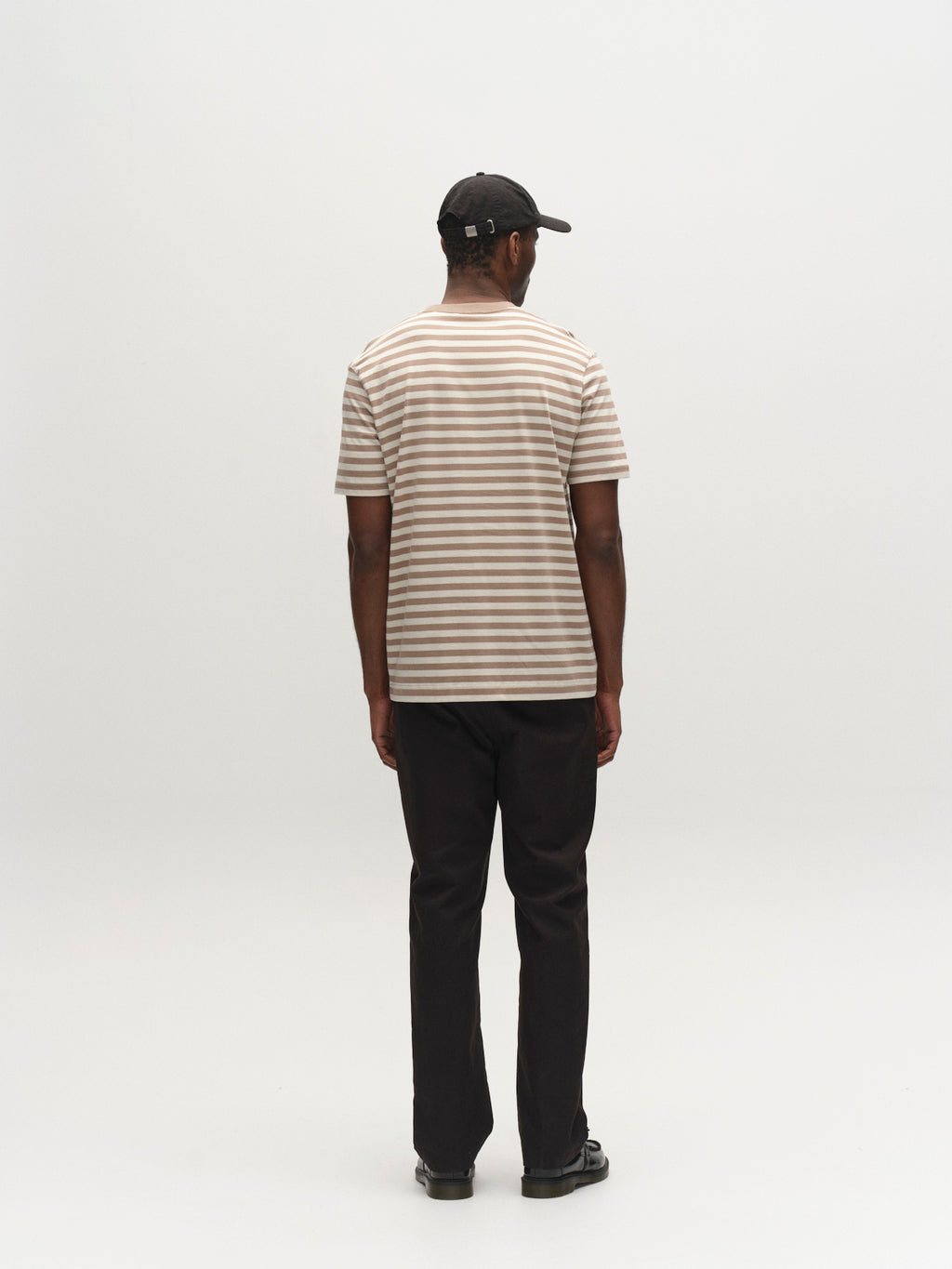 A person faces away from the camera, wearing a black cap, black pants and shoes, and the Gabba GABdune Logo Stripes SS GOTS - Fungi classic-fit beige and white organic cotton t-shirt against a plain white background.