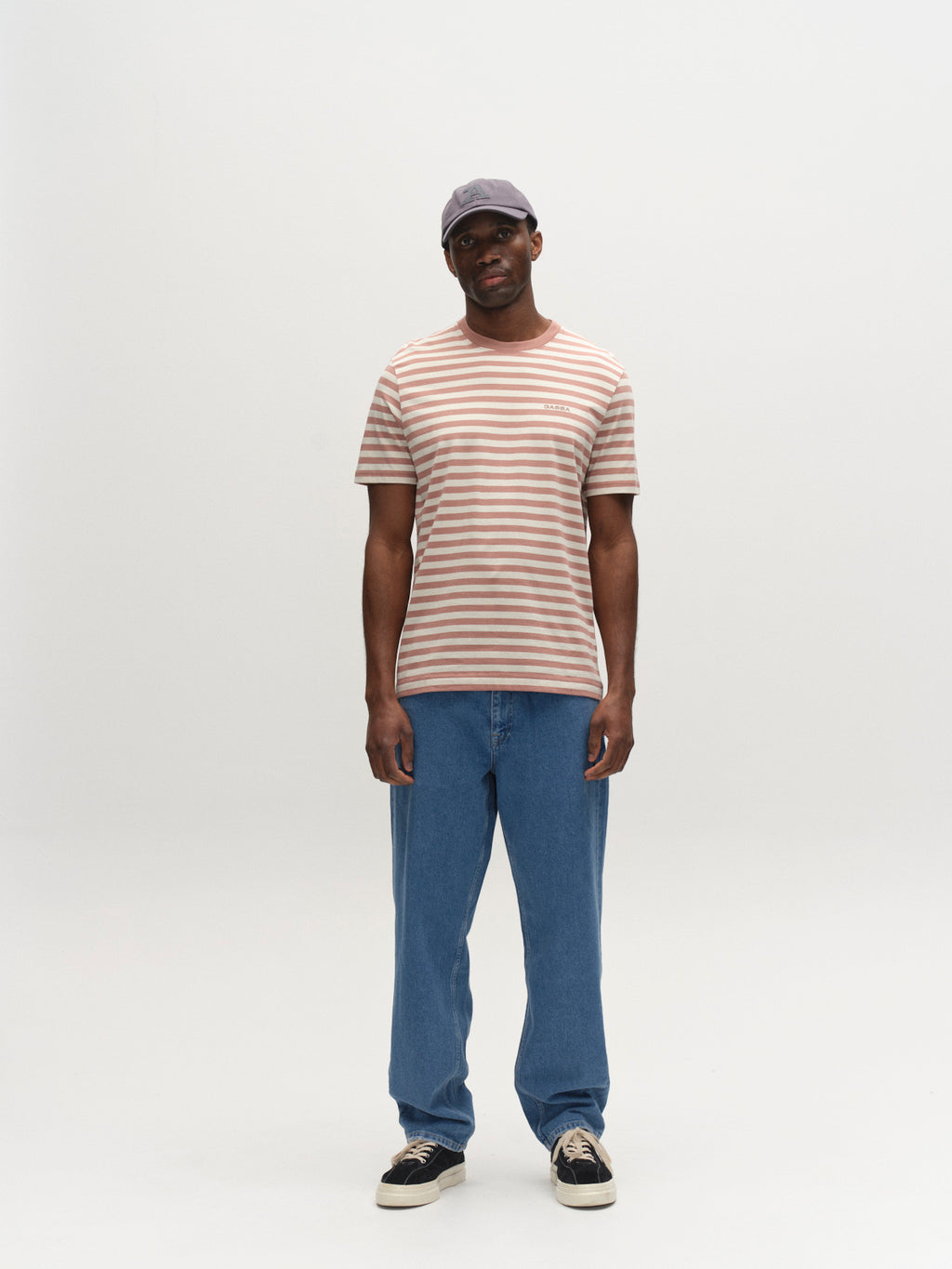 A man in a gray cap, blue jeans, black sneakers, and a Gabba GABdune Logo Stripes SS GOTS t-shirt in Wood Pink stands facing forward against a plain white background.