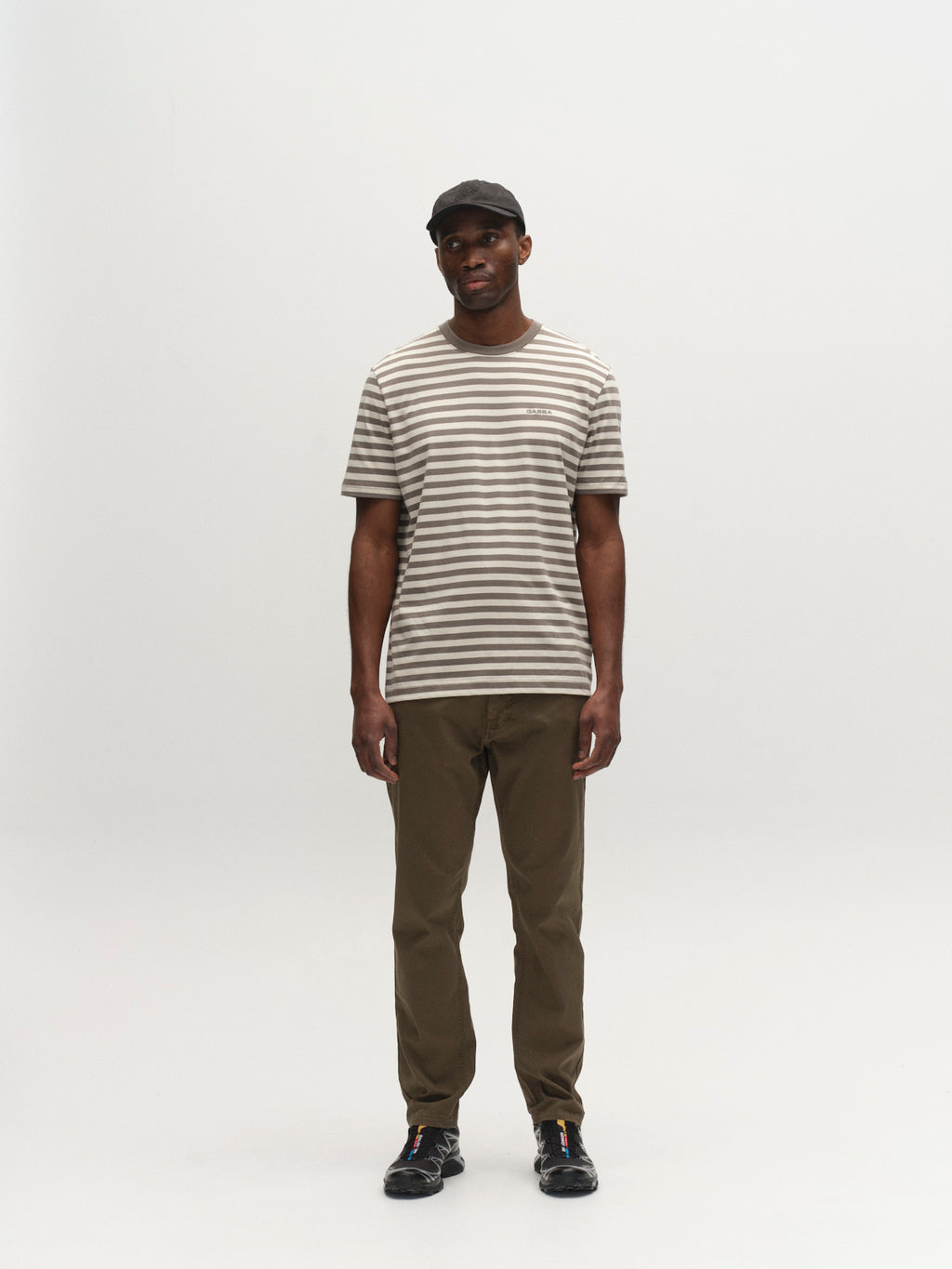 A man wearing a black cap, Gabba GABdune Logo Stripes SS GOTS - Castor Gray t-shirt, olive green pants, and black shoes stands facing forward against a plain white background.