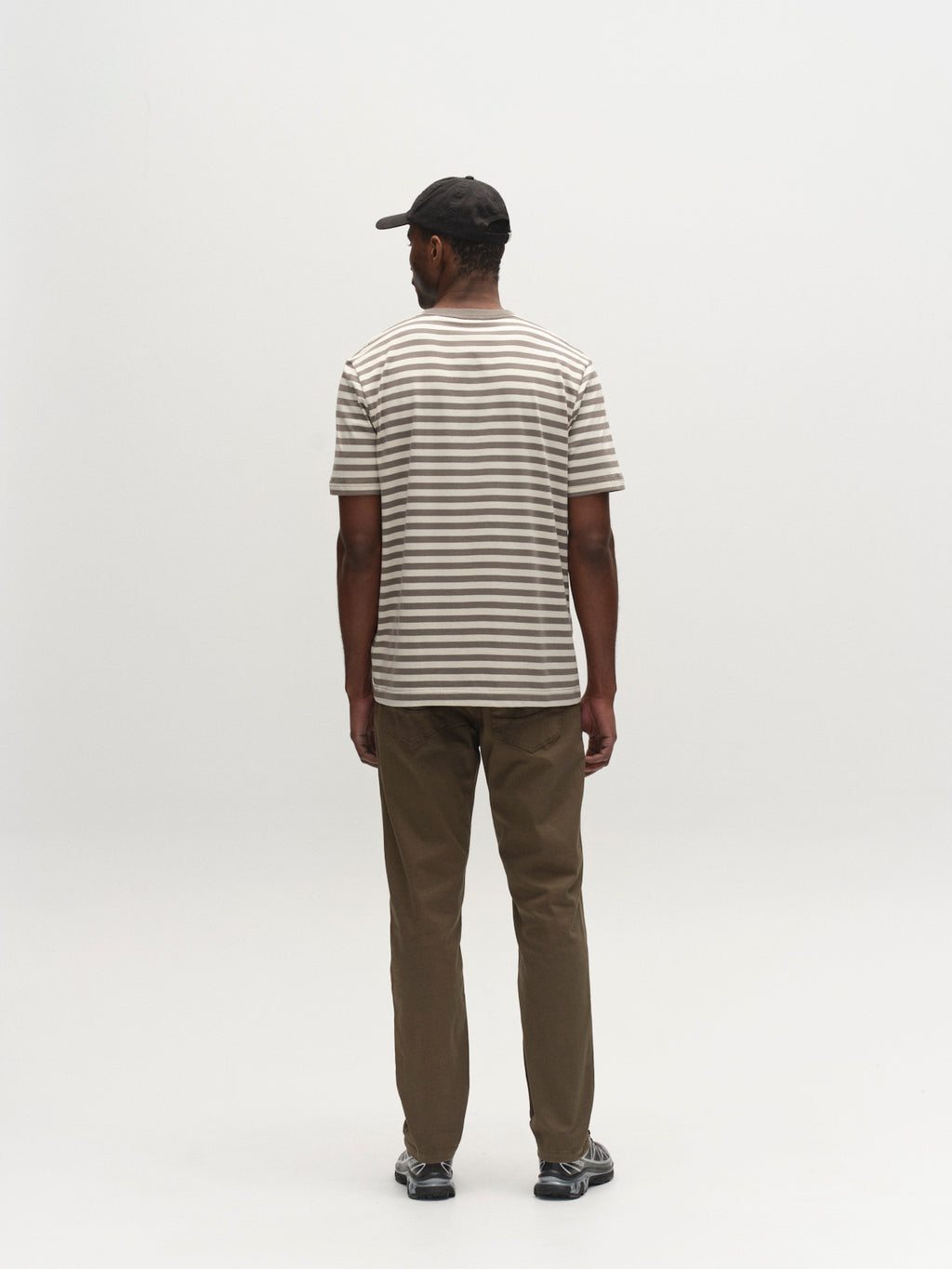 A man in a black cap, brown pants, black sneakers, and a Gabba GABdune Logo Stripes SS GOTS t-shirt in castor gray stands facing away in a minimal, light-colored studio.