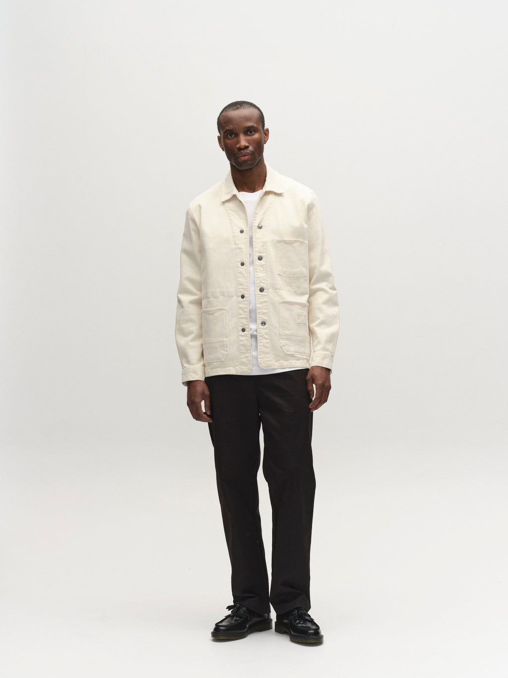 A man stands facing forward against a plain white background, wearing the Gabba GABbeta Twill - Ecru overshirt over a white shirt, paired with black pants and black shoes.