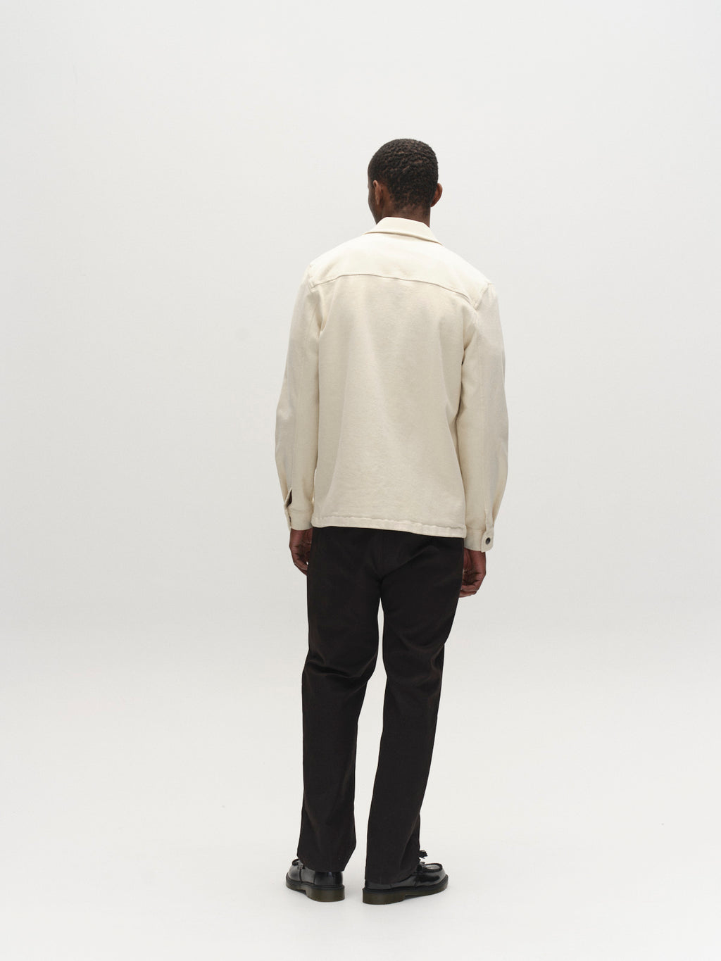 A man faces away from the camera against a white background, wearing the Gabba GABbeta Twill overshirt in ecru, paired with dark pants and black shoes.