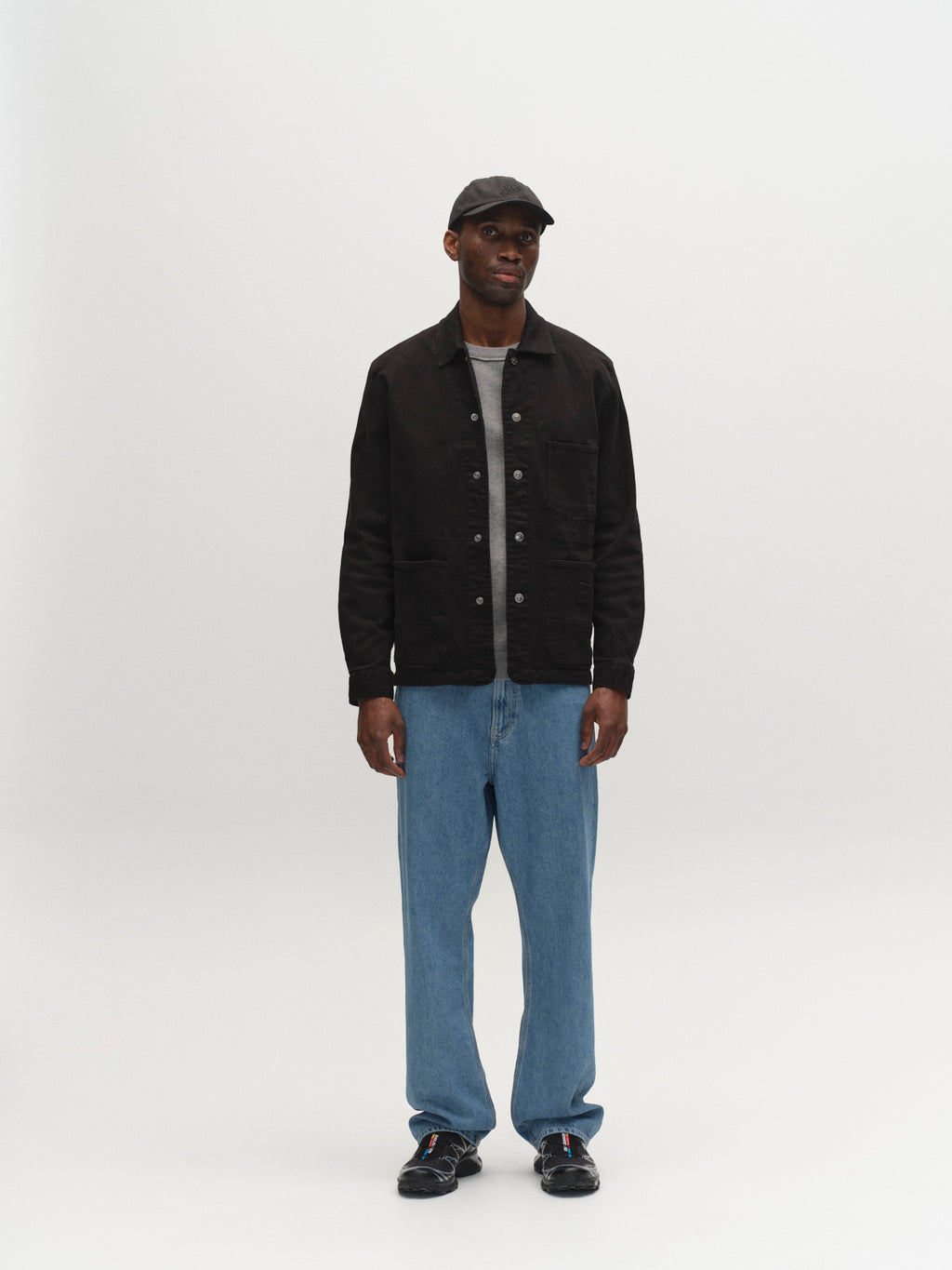 A person stands against a plain white background, wearing the Gabba GABbeta Twill - Black overshirt, a gray T-shirt, blue jeans, black sneakers, and a black baseball cap.