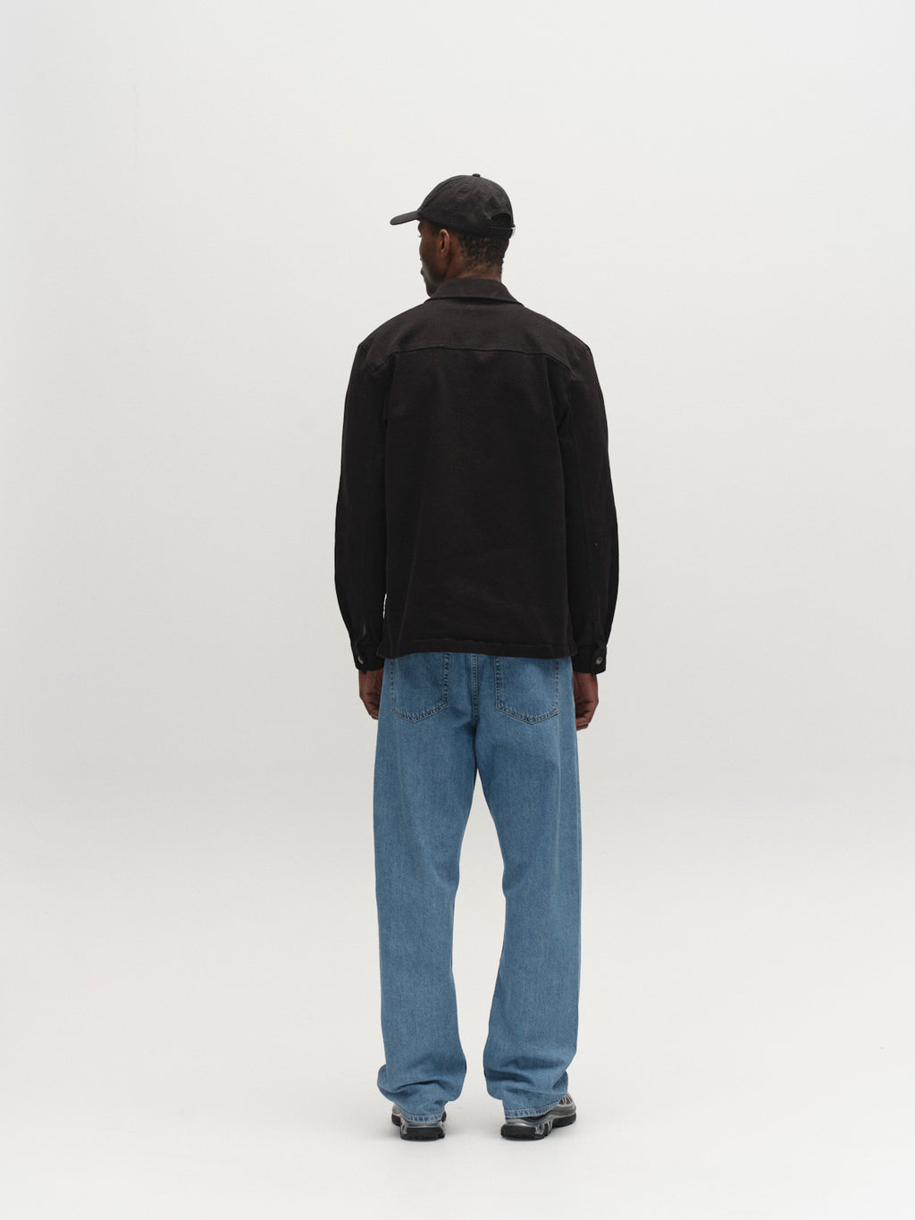 A man faces away from the camera against a white background, wearing Gabba's GABbeta Twill - Black overshirt, paired with blue jeans, black sneakers, and a black baseball cap.