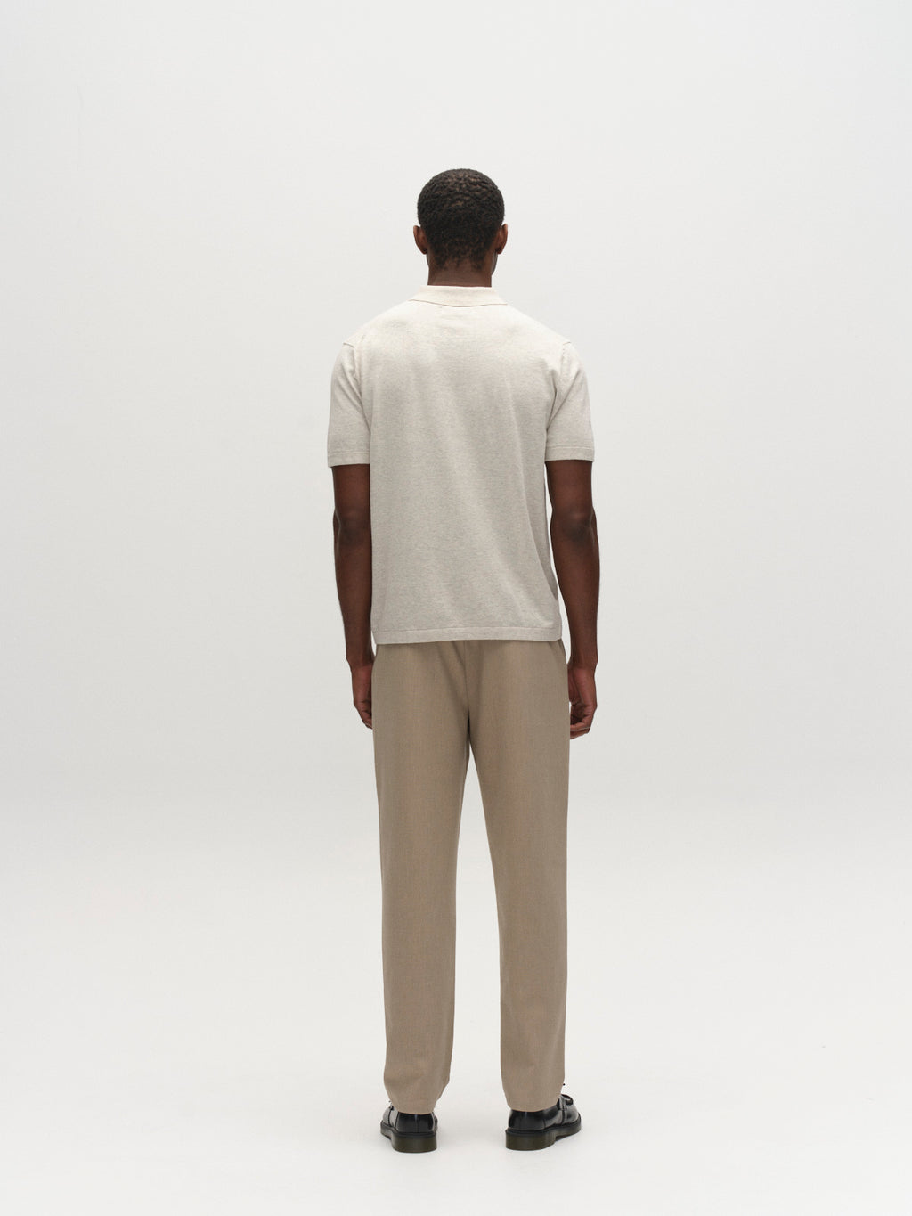 A man stands facing away in a plain room, wearing a light gray short-sleeve shirt, Gabba GABnapoli 19 Phin GRS - Sand Plaza chinos, and black shoes.