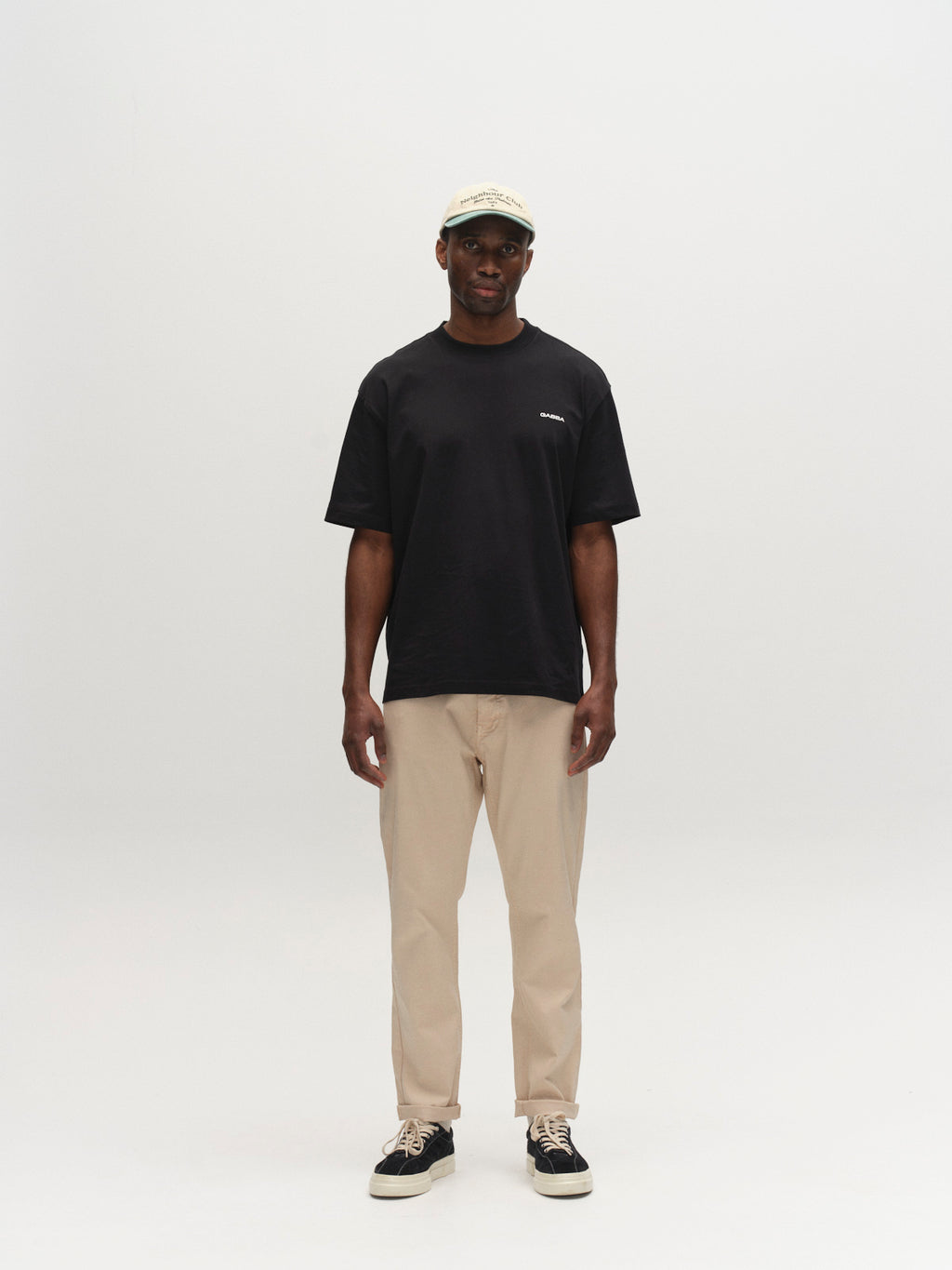 A man stands facing forward against a plain white background, wearing Gabba's GABmarc 5poc Marlo - Birch jeans, a beige cap, black t-shirt, and black sneakers.