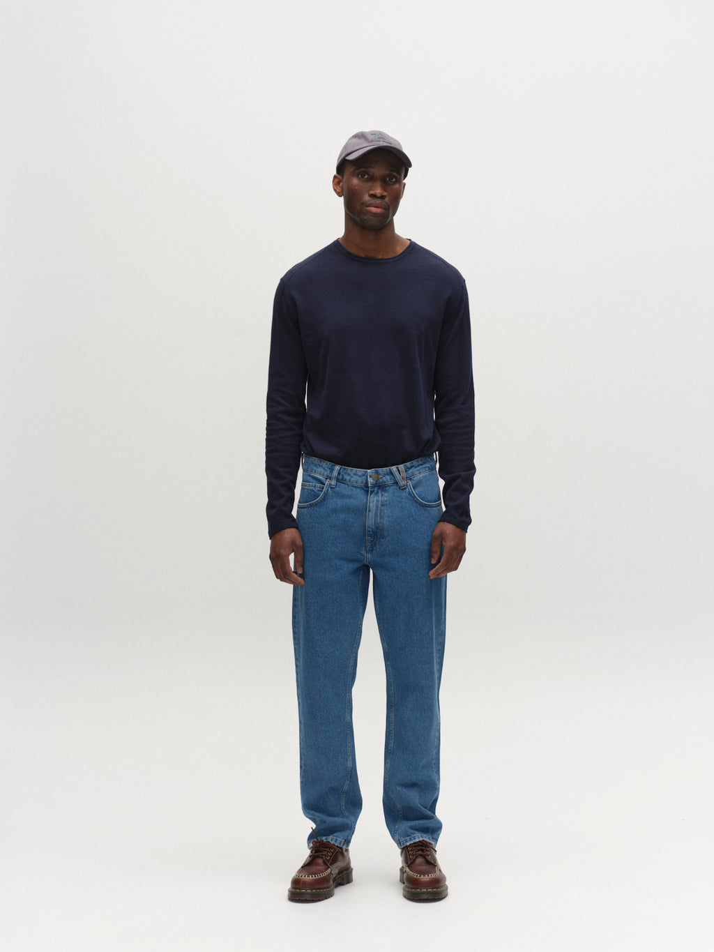 A man stands facing forward against a plain white background, wearing a navy long-sleeve shirt, Gabba GABmarc 5poc Mid Jeans in mid blue denim, brown shoes, and a black cap.