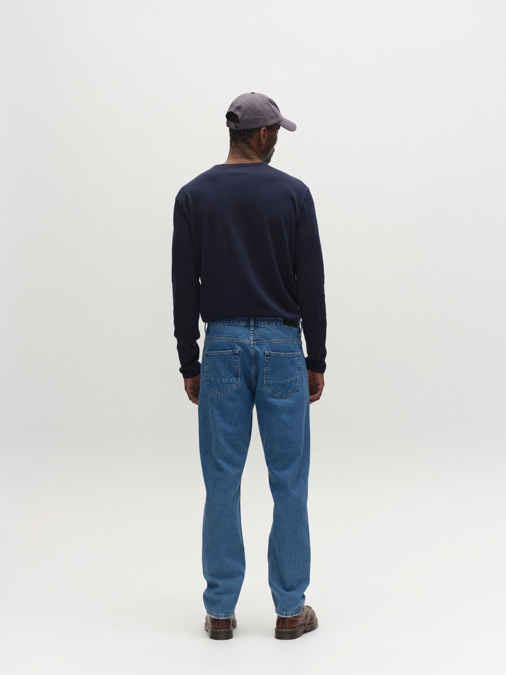 A person stands with their back to the camera, wearing a dark long-sleeve shirt, Gabba GABmarc 5poc Mid Jeans in mid blue denim, brown shoes, and a gray baseball cap against a plain white background.