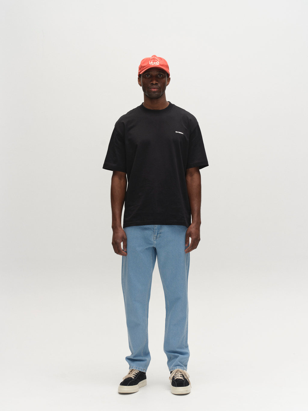 A person stands against a plain white background, wearing a black T-shirt, Gabba GABmarc 5poc Lig Jeans in Lt. Blue Denim, black-and-white sneakers, and a red baseball cap. They face forward with arms relaxed at their sides.