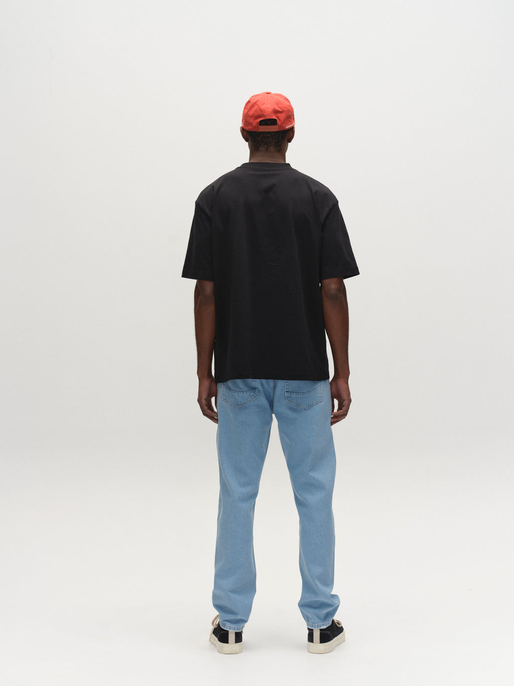 A man stands with his back to the camera, wearing a black T-shirt, Gabba GABmarc 5poc Lig Jeans in Lt. Blue Denim, black and white sneakers, and a red cap against a plain white background.