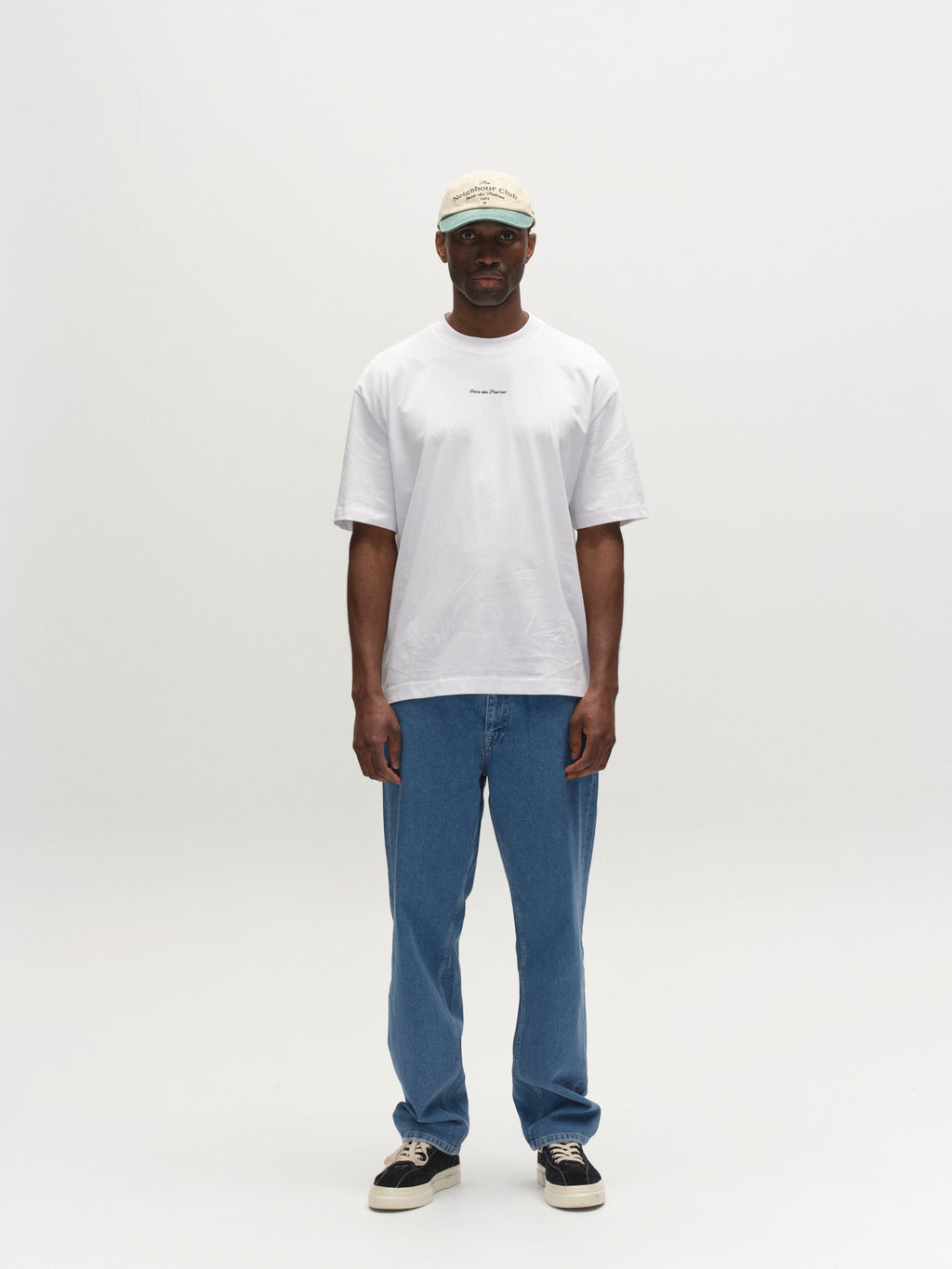A person faces forward against a plain white background, wearing a light-colored cap, white t-shirt, Gabba GABmax 5poc Mid Jeans in mid blue denim, and black sneakers.