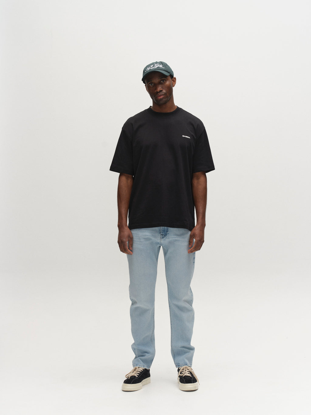 A person stands against a white background, wearing a dark green cap, black t-shirt, Gabba GABcarl Swif Jeans in light blue denim, and black sneakers with white soles. Their arms are relaxed by their sides.