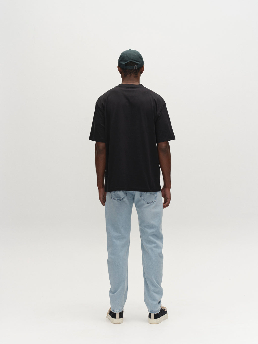 A person faces away from the camera, wearing a black t-shirt, Gabba GABcarl Swif Jeans in Lt. Blue Denim, black and white sneakers, and a dark green cap against a plain white background.