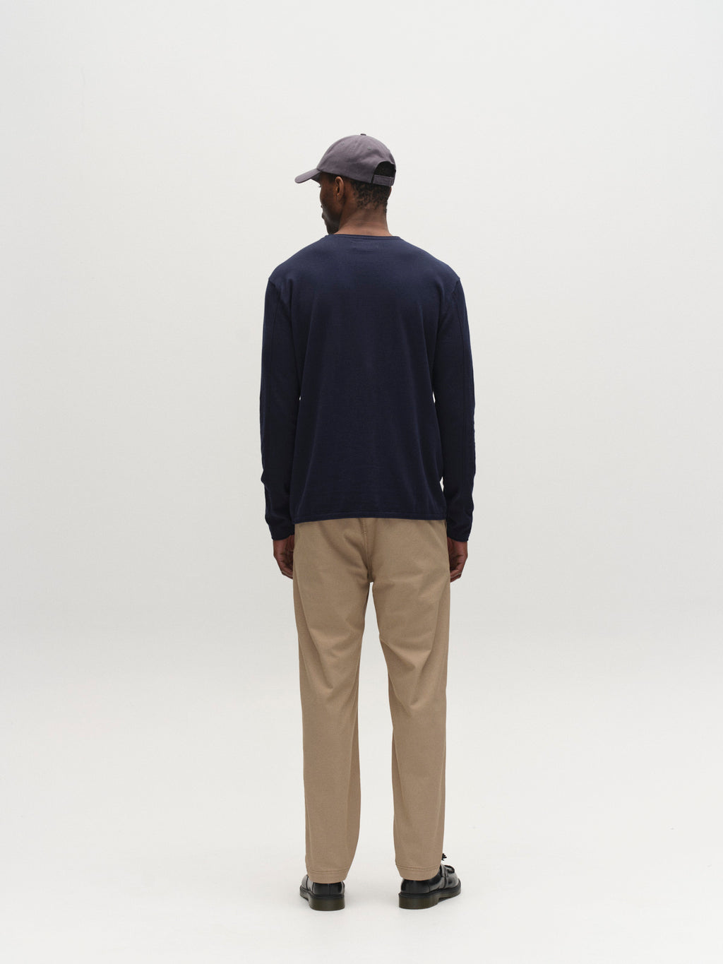 A man faces away from the camera against a white background, wearing a navy long-sleeve shirt, black shoes, a grey cap, and Gabba GABjoe Nopleats Dale Pant in Sand—a modern tapered-fit chino in high-stretch cotton-blend fabric.