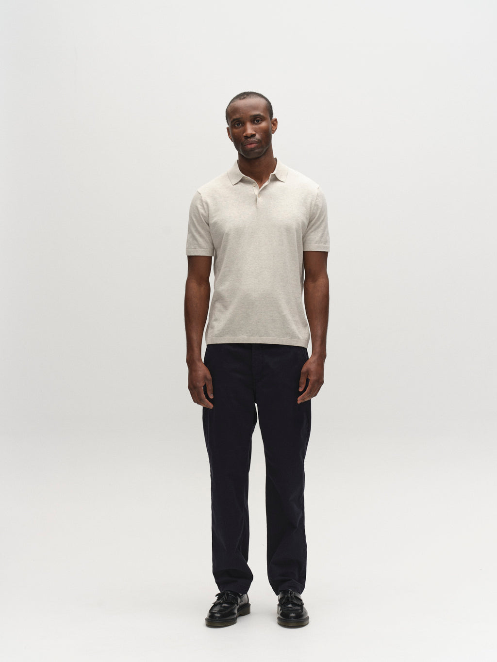 A man stands against a plain white background, facing forward in a light beige polo and Gabba GABjoe Nopleats Dale Pant - Blue Black, paired with black shoes. His neutral expression and relaxed posture showcase this modern wardrobe essential.