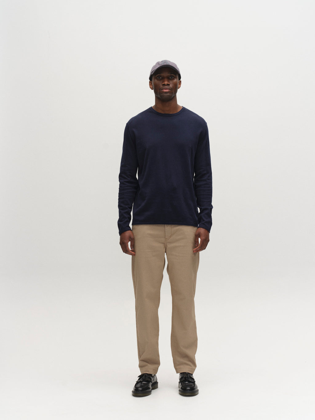 A person faces forward against a plain white background, wearing the Gabba GABlima Cot LS Knit navy long-sleeve shirt, beige pants, black shoes, and a dark baseball cap.