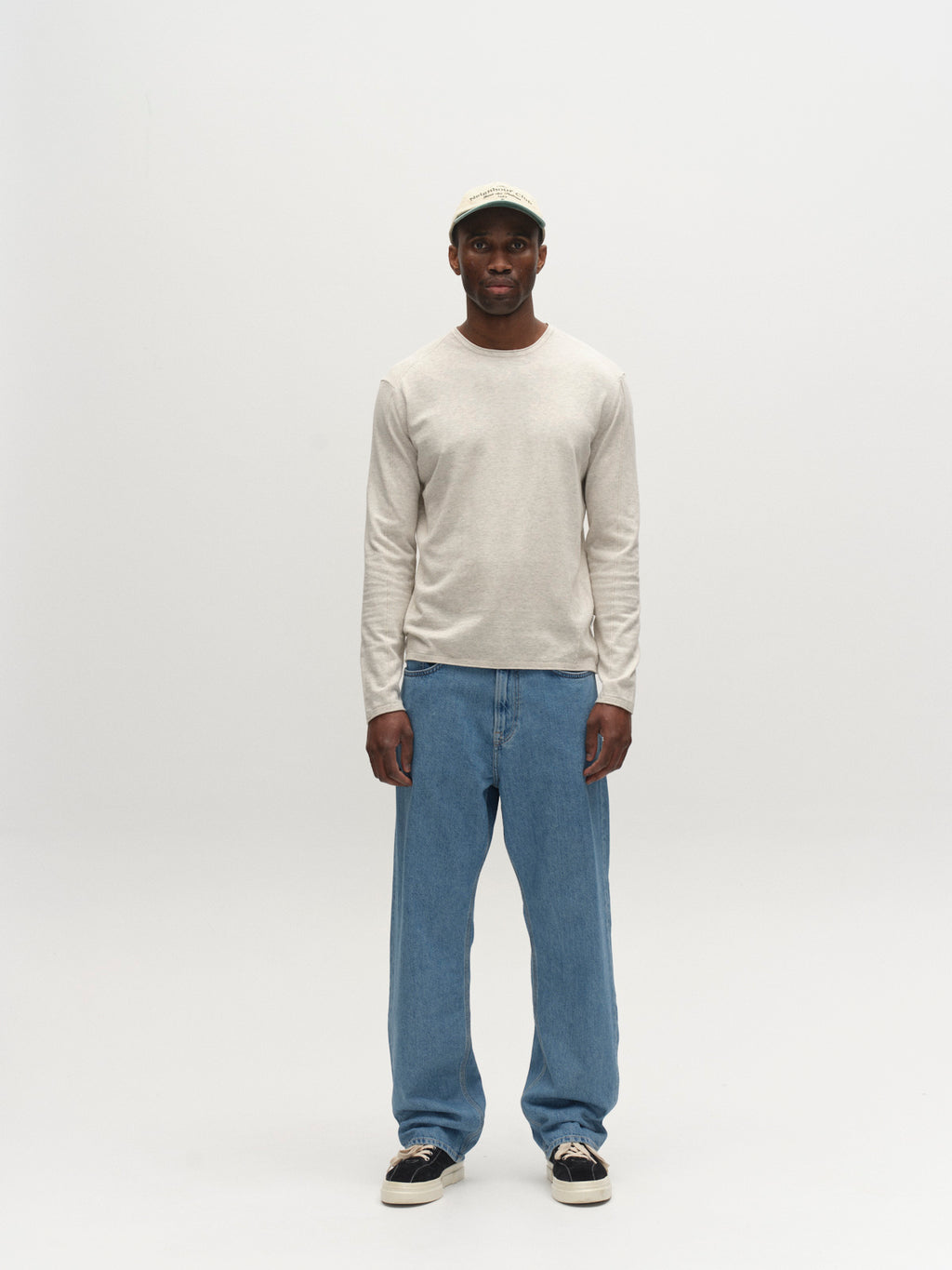 A man stands against a white background wearing the Gabba GABlima Cot LS Knit in Sand Mel. He pairs it with loose blue jeans, black sneakers, and a light-colored baseball cap, facing forward with relaxed arms at his sides.
