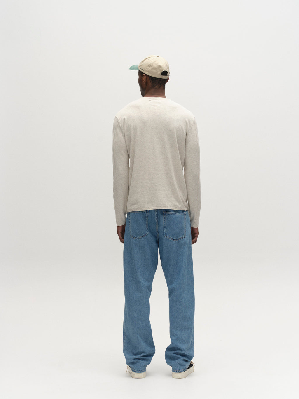 A person wears the Gabba GABlima Cot LS Knit - Sand Mel, paired with loose blue jeans, white sneakers, and a beige baseball cap, standing facing away in a plain, light-colored studio setting.