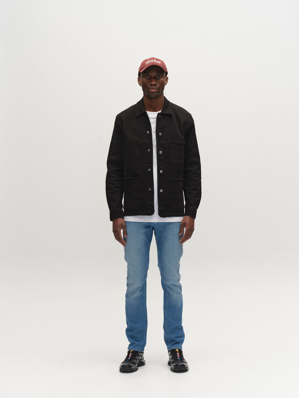 A man stands facing forward against a plain white background, wearing a red cap, black jacket, white t-shirt, Gabba GABjones Mik Jeans in Lt. Blue Denim, and black shoes.