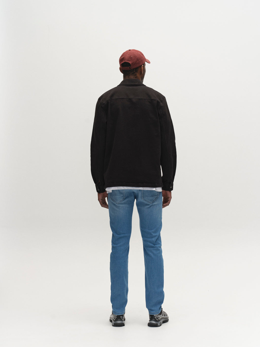 A person faces away from the camera in a red cap, black jacket, sneakers, and Gabba GABjones Mik Jeans in Lt. Blue Denim against a plain white background.