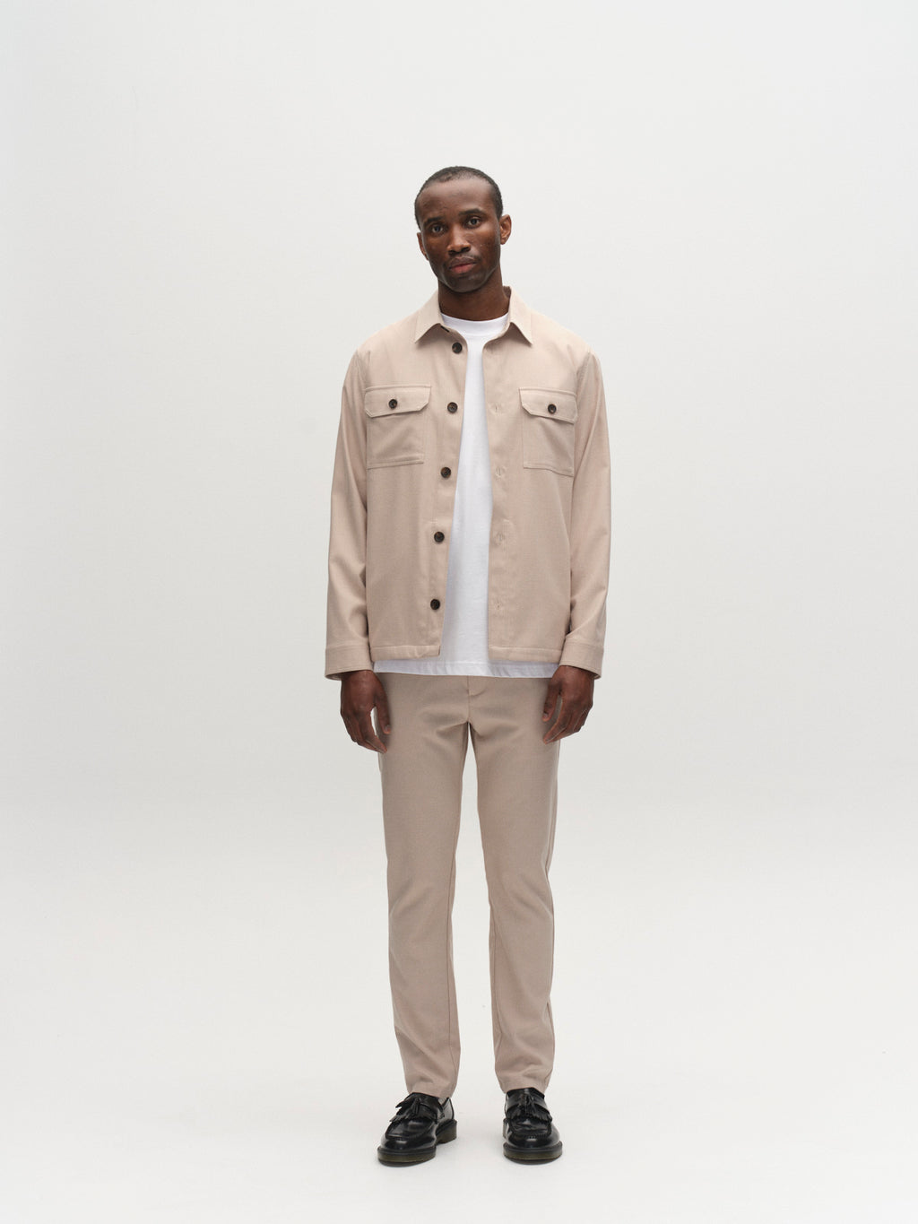 A man stands against a white background, wearing Gabba's GABpaul Deme Pant in Birch with a beige jacket over a white t-shirt and black shoes. His arms rest at his sides and he has a neutral expression.