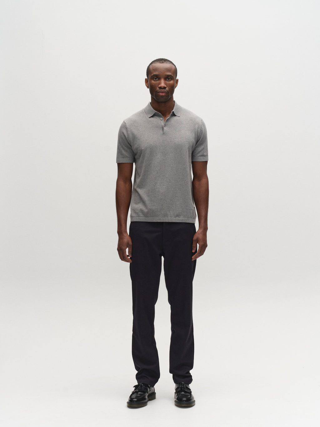 A man stands facing forward against a plain white background, wearing a short-sleeve gray polo shirt, the Gabba GABpaul Deme Pant in dark navy featuring a slim chino fit and micro-pattern woven fabric, along with black shoes—an ideal look for everyday wear.