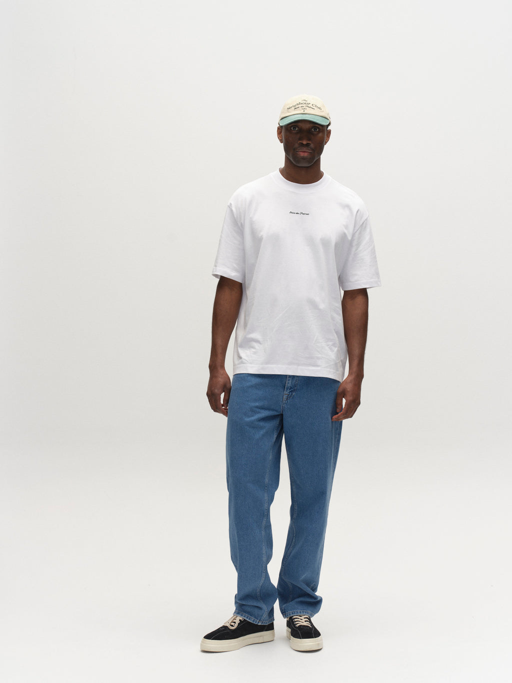 A man stands against a plain white background wearing a Gabba GABnigel NBH SS T-Shirt GOTS in white, blue jeans, black and white sneakers, and a light-colored baseball cap.