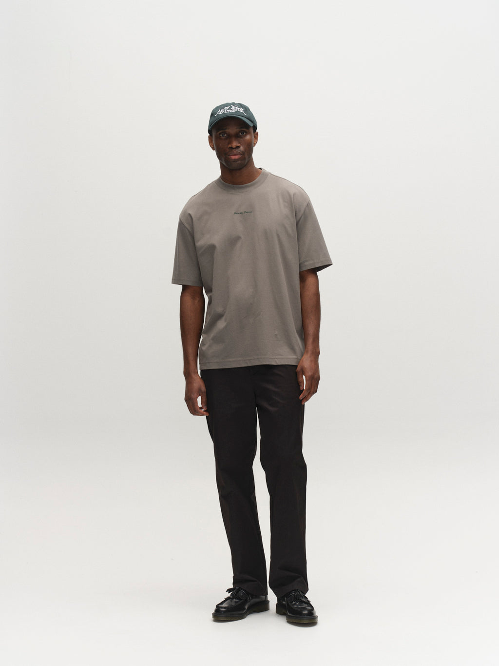 A man with a neutral expression stands against a white background, wearing a dark green cap, black pants, black shoes, and the Gabba GABnigel NBH SS T-Shirt GOTS in Castor Gray.