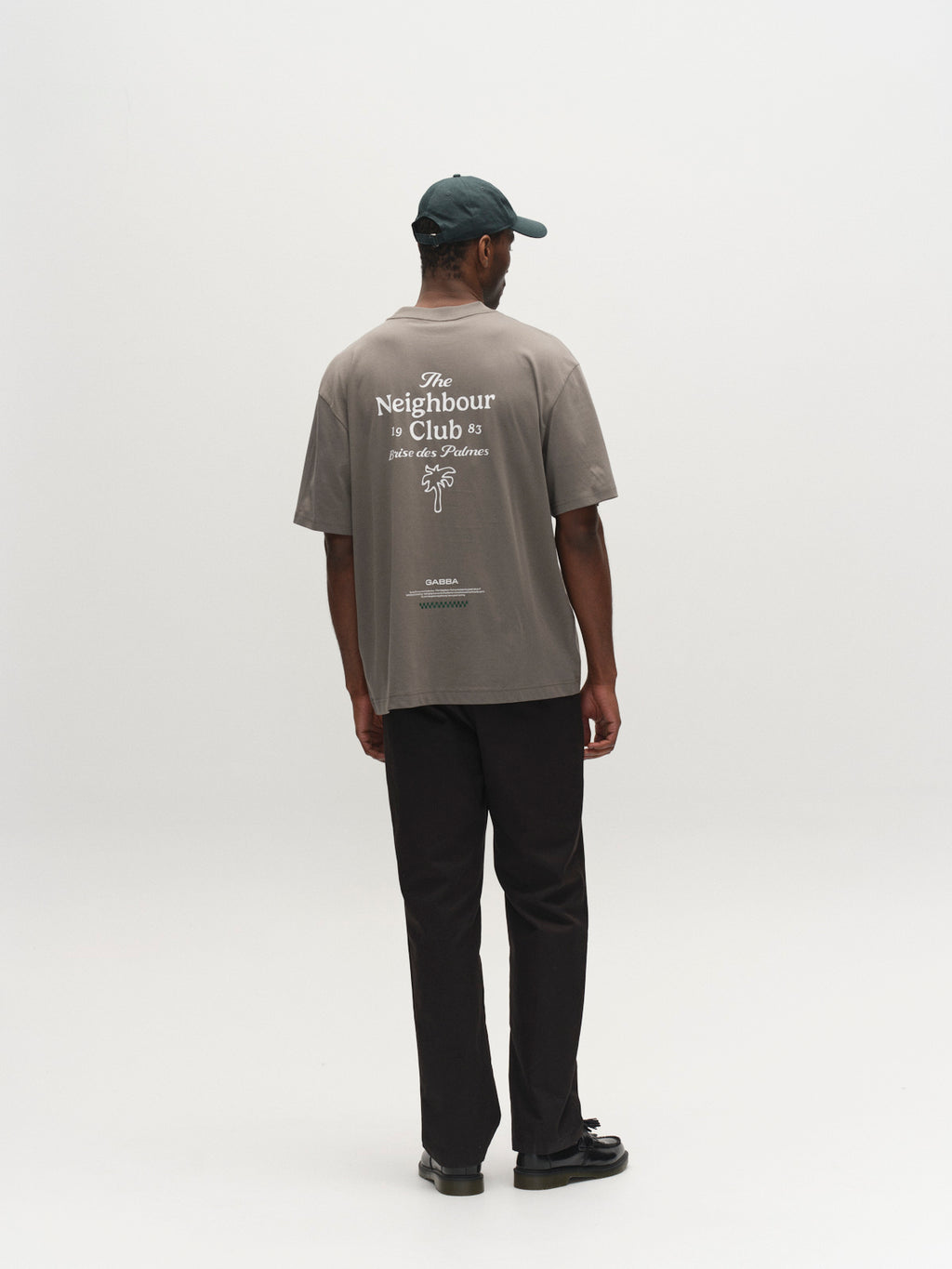 A person wears a Gabba GABnigel NBH SS T-Shirt GOTS in Castor Gray and black pants, standing with their back to the camera in a minimal white studio, showcasing the shirt’s white text and graphics.