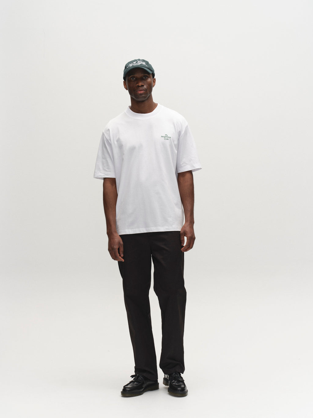 A man stands against a white background wearing the Gabba GABnigel Boxy NBH SS T-shirt GOTS in white, paired with black pants, black shoes, and a dark baseball cap. He faces the camera with a relaxed posture and neutral expression.