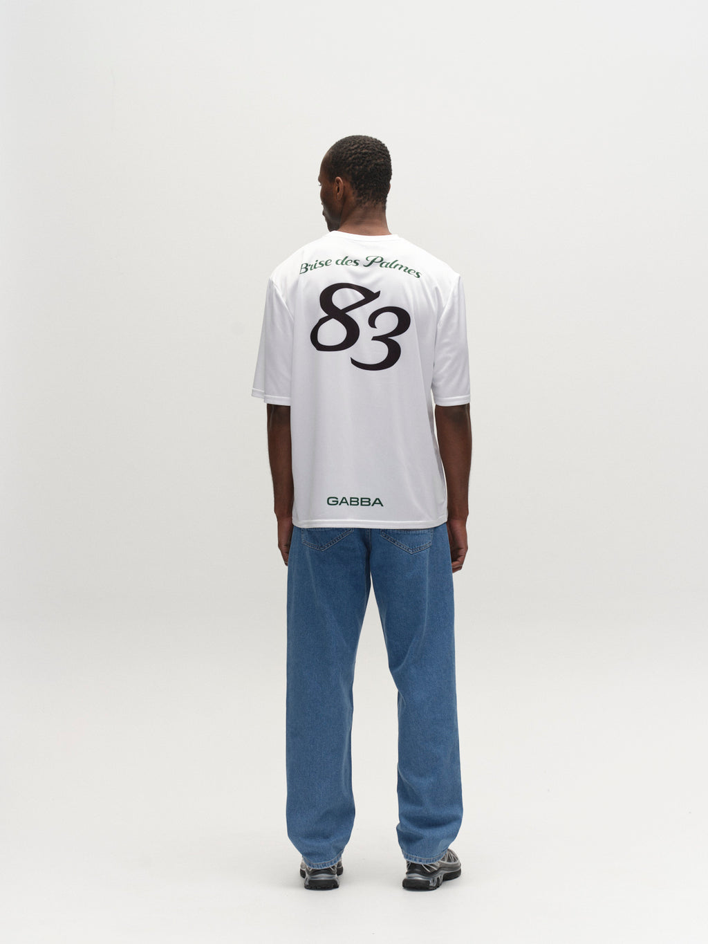 A man faces away in a Gabba GABnigel Fut Boxy Pes SS T-shirt - White, featuring Brise des Fleurs and a bold 83 print on the back, styled with blue jeans and sneakers against a plain light background.