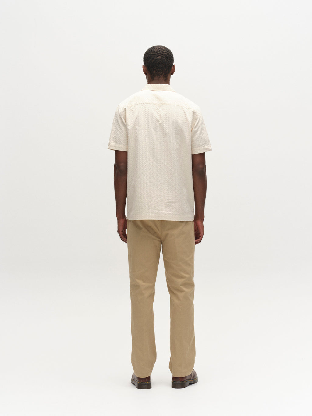 A man stands with his back to the camera wearing a short-sleeved off-white shirt, dark shoes, and Gabba's GABdenver Fuggies Pant in Sand against a plain, light background.