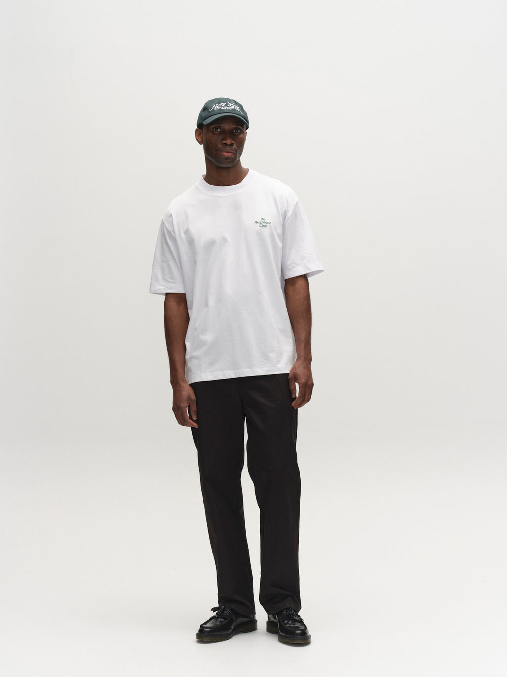 A person stands against a plain white background, wearing a white t-shirt, black shoes, dark baseball cap and glasses, and Gabba GABdenver Fuggies Pant in Black.