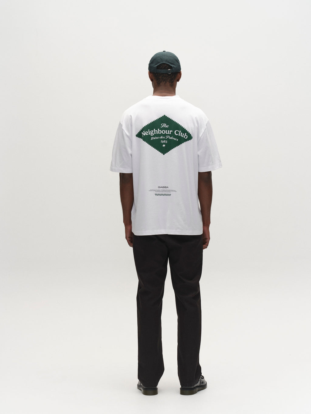 A person stands facing away, wearing a dark green cap, a white tee with 