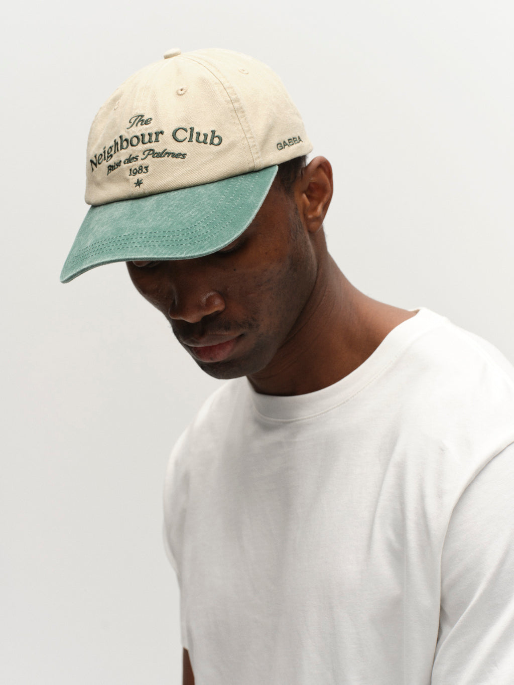 A man in a Gabba GABnbh Club Cap - Green, featuring The Neighbour Club on the front and crafted from soft material, paired with a plain white t-shirt, looks down against a simple light background.