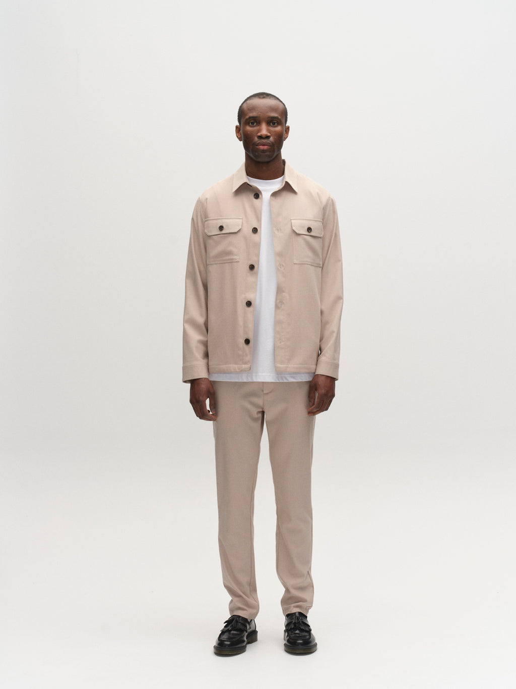 A man stands facing forward against a plain white background, wearing the Gabba GABtogger Base Deme LS Overshirt in Birch with matching beige pants, a white T-shirt, and black shoes—ideal for everyday wear or style tests.