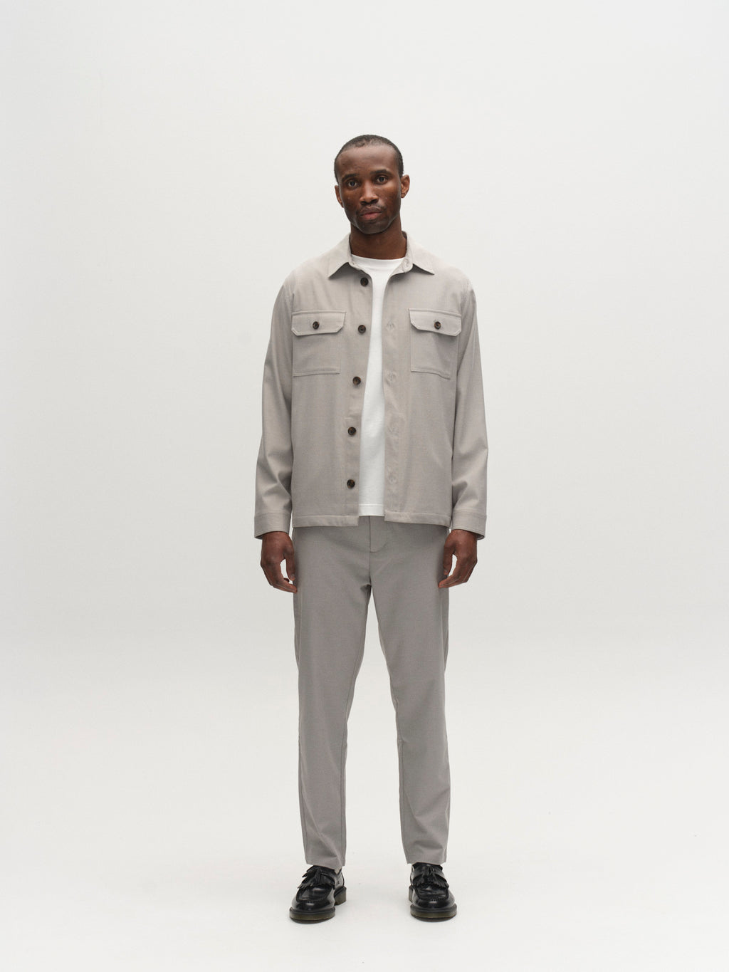 A man stands facing forward against a white background, wearing the Gabba GABtogger Base Deme LS Overshirt in Mineral Gray with matching gray pants, a white shirt, and black shoes. He has a neutral expression and his arms at his sides.