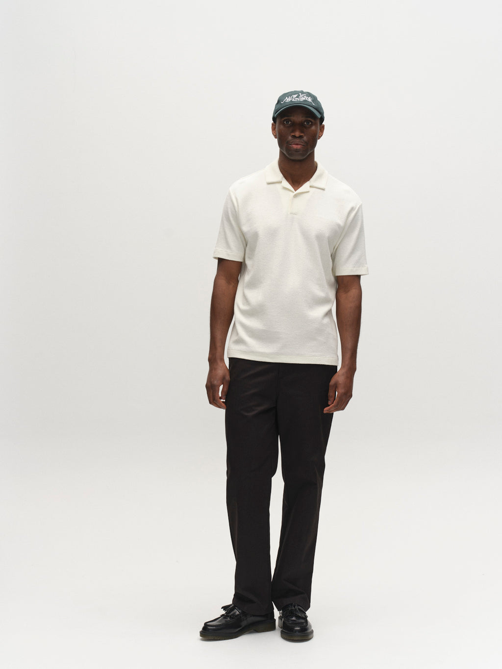 A man faces forward with a neutral expression, wearing Gabba's GABlio Sku Towel T-Shirt in Egret, paired with black pants, black shoes, and a dark green baseball cap against a plain white background.
