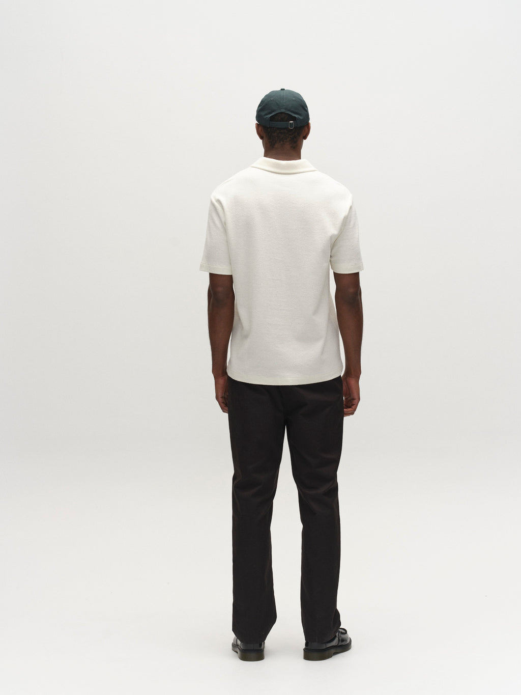 A person wearing the Gabba GABlio Sku Towel T-Shirt - Egret, black pants, dark shoes, and a dark baseball cap stands with their back to the camera against a plain white background.