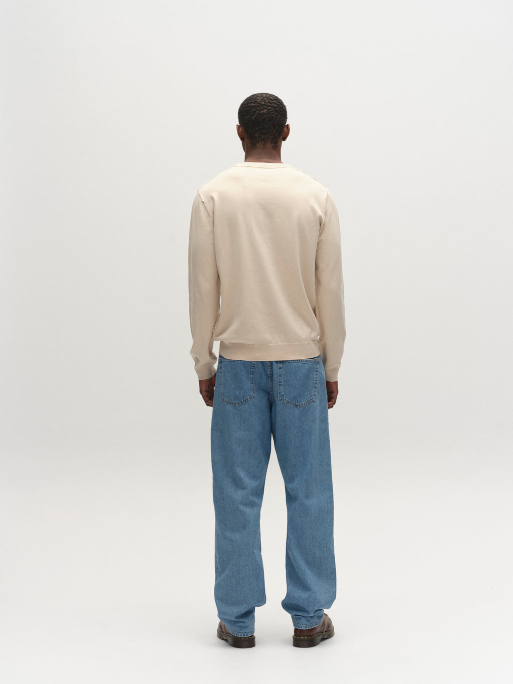 A person faces away from the camera against a plain white background, wearing the Gabba GABfumo Spy LS Knit in sand, loose blue jeans, and brown shoes.